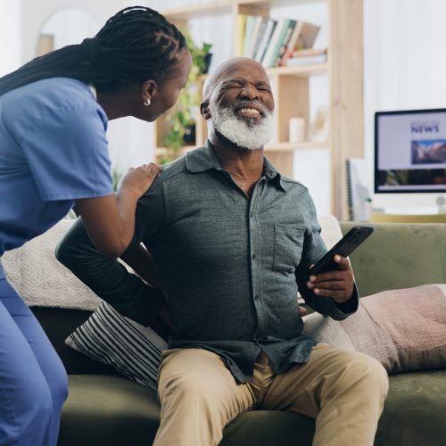 Nurse, senior man and back pain on sofa for help, care or support for recovery from injury in retirement. Black people, physical therapy and elderly person with muscle, rehabilitation and watching tv.